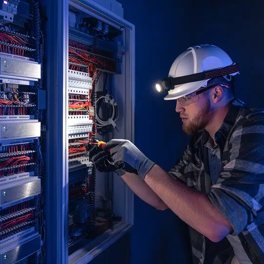 electrician with white hard hat, head lamp and clear safety goggles repairing electrical wiring in panel