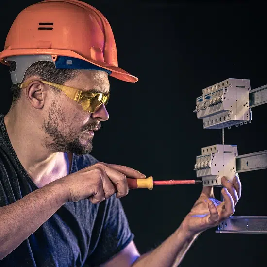 electrician with orange hard hat and yellow safety glasses holding screwdriver up to electrical outlet and switch box